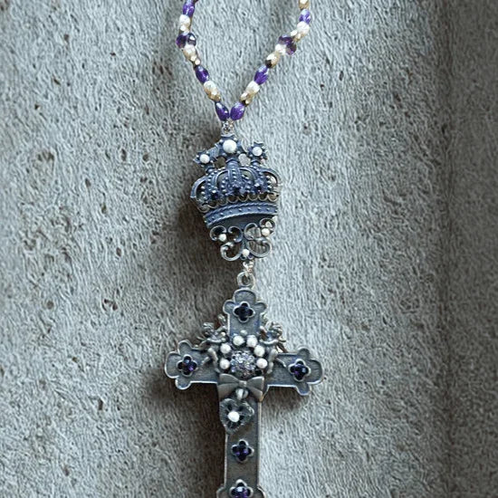 Jeweled silver cross with crown, purple bead necklace on textured background